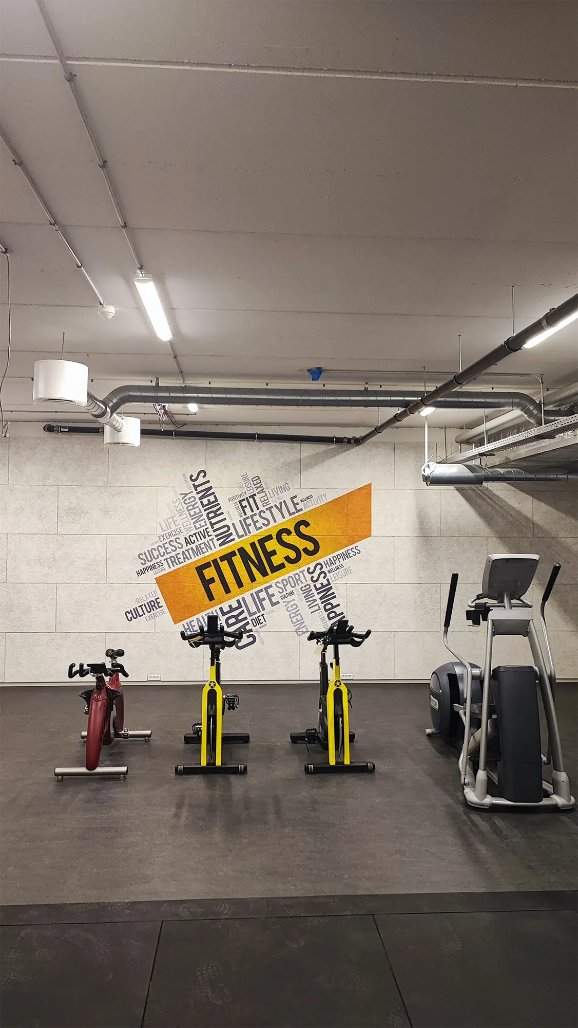 Træningsrum med spinningcykler og crosstrainer foran en væg printet med en ordsky og gult FITNESS-felt