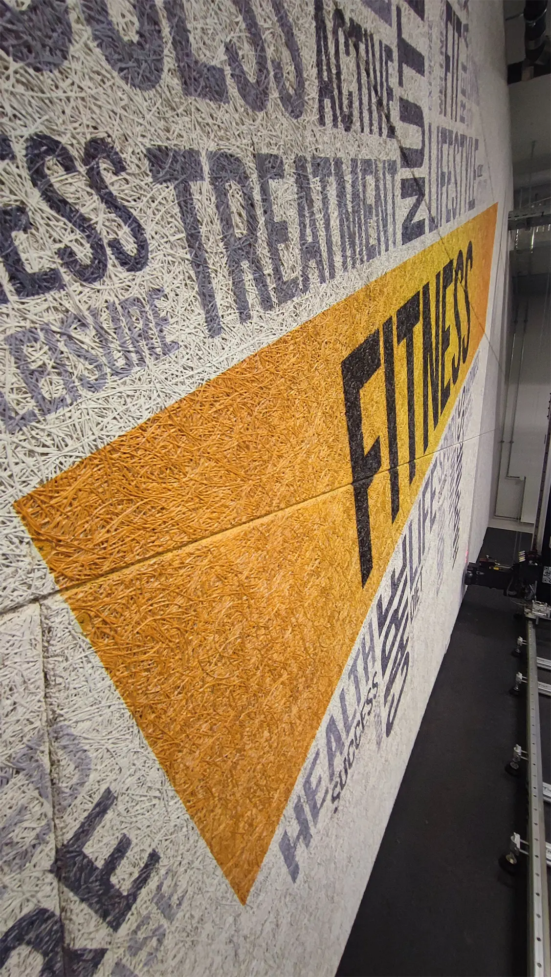 Skråt nærbillede af det printede FITNESS-motiv, hvor trykket følger vægoverfladens struktur