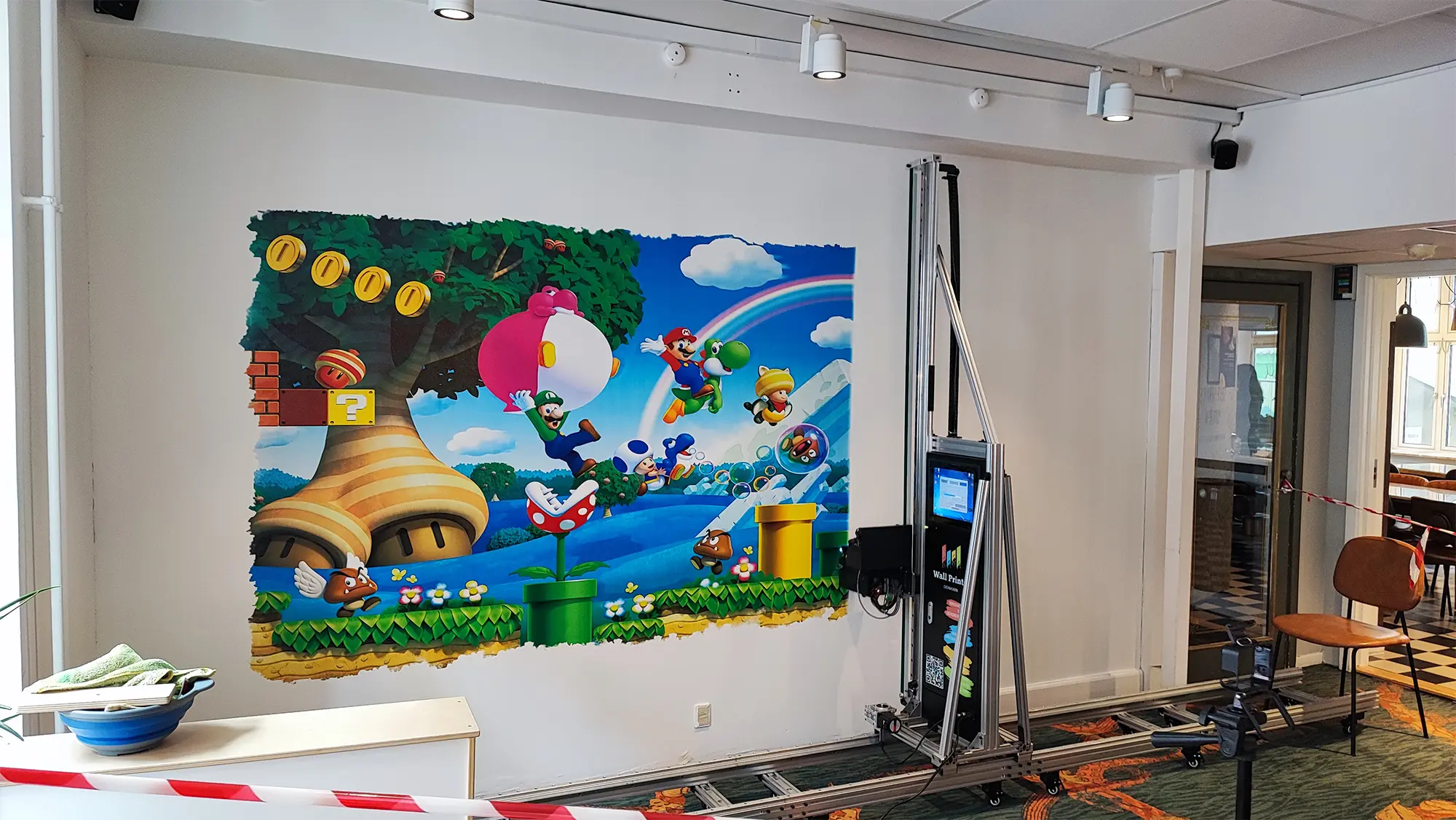 Vægprinteren opstillet på skinner foran gaming-motivet under arbejdet i fællesarealet