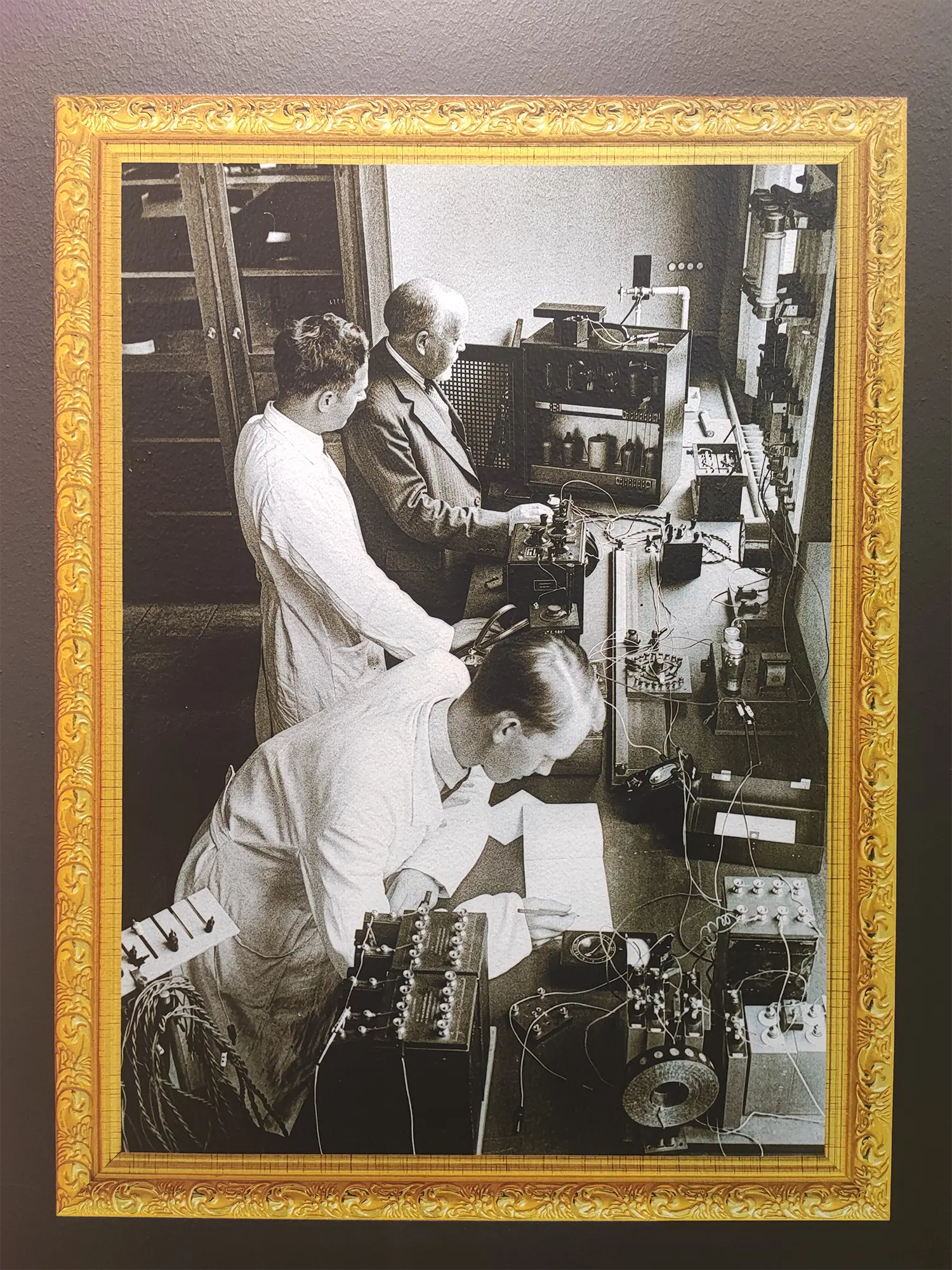 Historisk laboratoriefoto med tre medarbejdere ved måleudstyr, printet på væggen i stående format